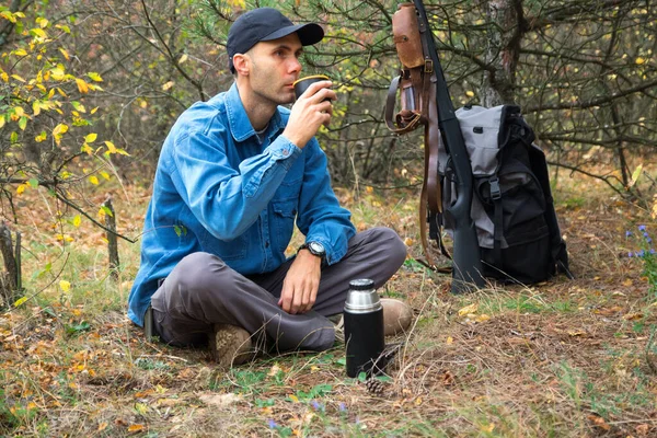 白人男性ハンターが森で休んでいます。ポットからお茶を飲みます。散弾銃、弾薬、背景にバックパック — ストック写真