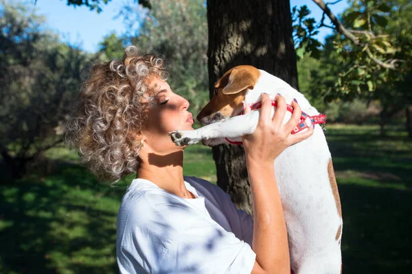 Blondynka kręcone dziewczyna trzyma na ręce i całuje jej zwierzak, Jack Russell pies w jesiennym parku — Zdjęcie stockowe