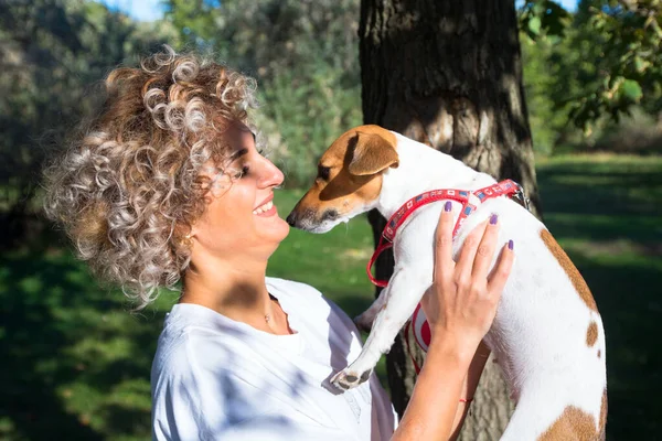 Blondynka kręcone dziewczyna trzyma na ręce i całuje jej zwierzak, Jack Russell pies w jesiennym parku — Zdjęcie stockowe