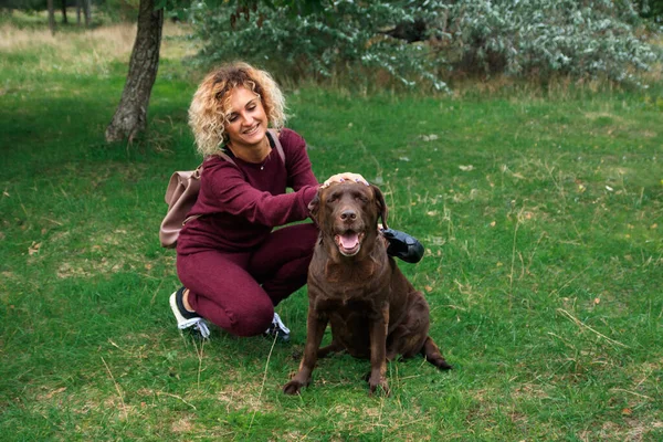 Blondynka kręcone dziewczyna bawi się z czekolady labrador w zielony park — Zdjęcie stockowe