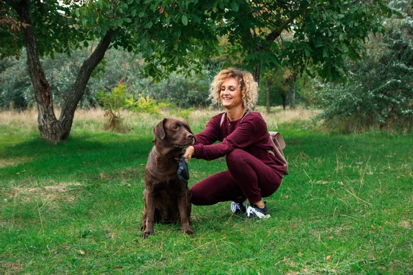 Blondynka kręcone dziewczyna bawi się z czekolady labrador w zielony park — Zdjęcie stockowe