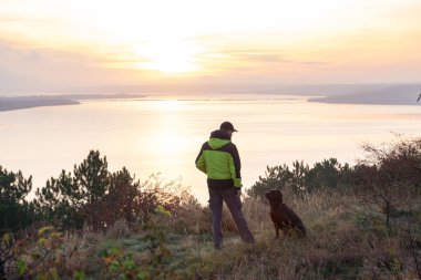 Labrador köpeğiyle gölün üzerinde turuncu gündoğumuna hayran olan bir adam.