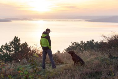 Labrador köpeğiyle gölün üzerinde turuncu gündoğumuna hayran olan bir adam.