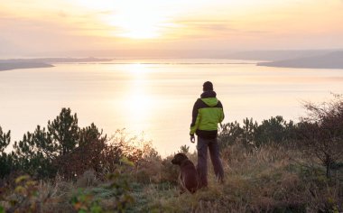 Labrador köpeğiyle gölün üzerinde turuncu gündoğumuna hayran olan bir adam.