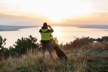 Labrador köpeğiyle gölün üzerinde turuncu gündoğumuna hayran olan bir adam.
