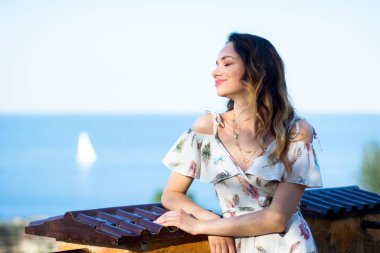 Terasdaki beyaz elbiseli kız deniz ve yat manzarasının tadını çıkarıyor.