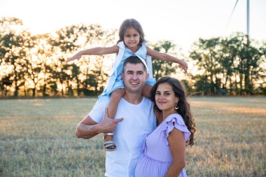 Mutlu bir koca, hamile bir eş ve babasının omuzlarında küçük bir kız. Arka planda rüzgar türbinleri.