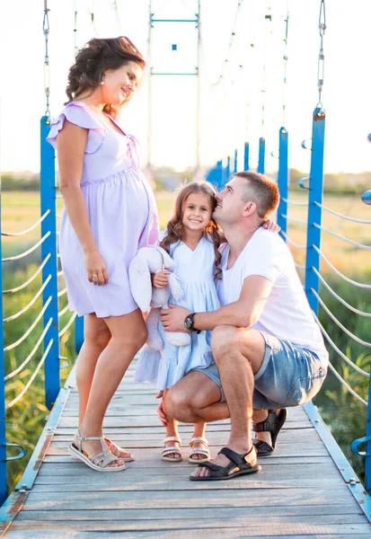 happy young family are hugging on the bridge in the sunset light. husband and a little girl touching pregnant wife\'s belly