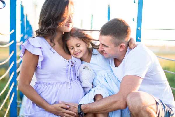 Mutlu genç aile gün batımında köprüde sarılıyor. Koca ve küçük bir kız hamile kadının karnına dokunuyor.