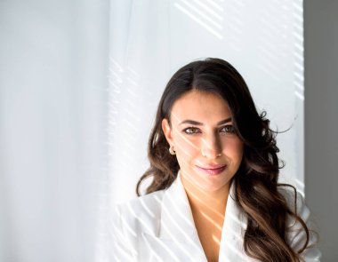 Beyaz ceketli, güzel gözleri ve Yunan yüz hatları olan beyaz bir kadın. Pencere kenarında külüstürle oturuyor. Yüzünde ve ceketinde güneş ışığı çizgileri var.