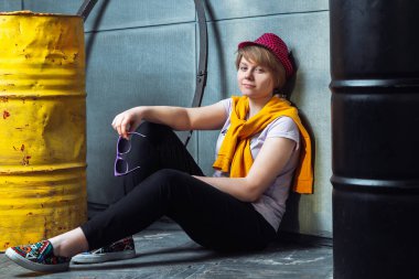 Kız iki varil yanındaki duvarın yanında yerde oturuyor.