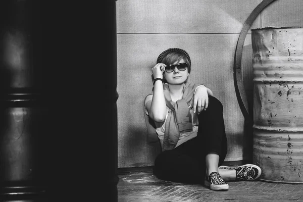 The girl is sitting on the floor near the wall next to two barrels