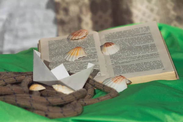 Een open boek met schelpen liggen naast het touw en een papieren boot — Stockfoto