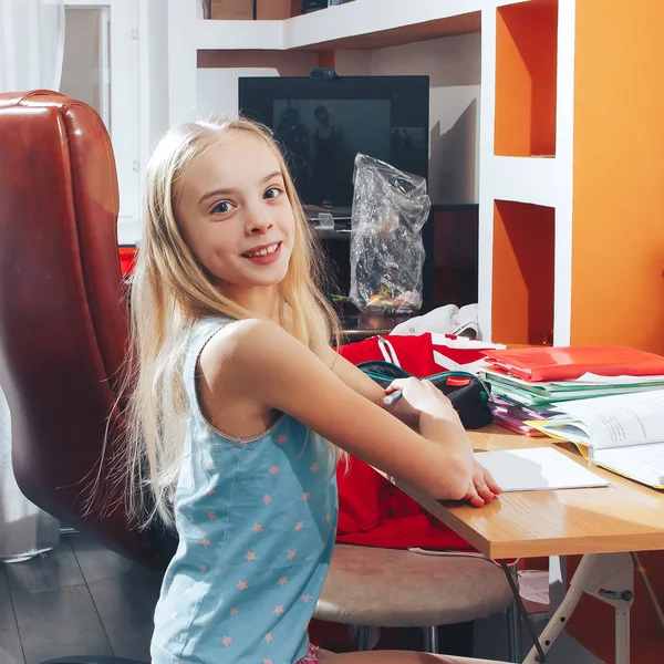 Hermosa niña rubia se sienta a la mesa y hace los deberes — Foto de Stock