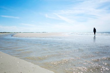 Kumlu tropikal plajda gelgit bölgesinde dalgalanan sığ deniz suyu ve su yüzeyinde kısmi yansıması olan uzak insan silueti. Deniz manzarası düşük açılı. Yaz tatili kavramı