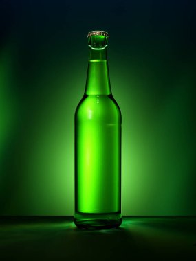 Etiketsiz tek yeşil cam bira şişesi. Yeşil ışıklandırılmış arka planda metal kapaklı dolu bira şişesi. Alkolik içki konsepti.