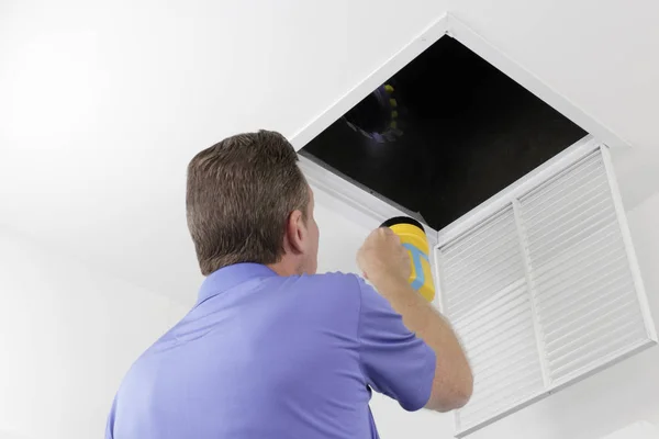 Adam bir hava kanalı bir el feneri ile teftiş. Hvac kanalları büyük bir kare delik içinde inceleyerek sarı bir el feneri ile büyük bir erkek. Erkek teknisyen bir ev hava girişi deliği içinde hava kanalları üzerinde seyir.