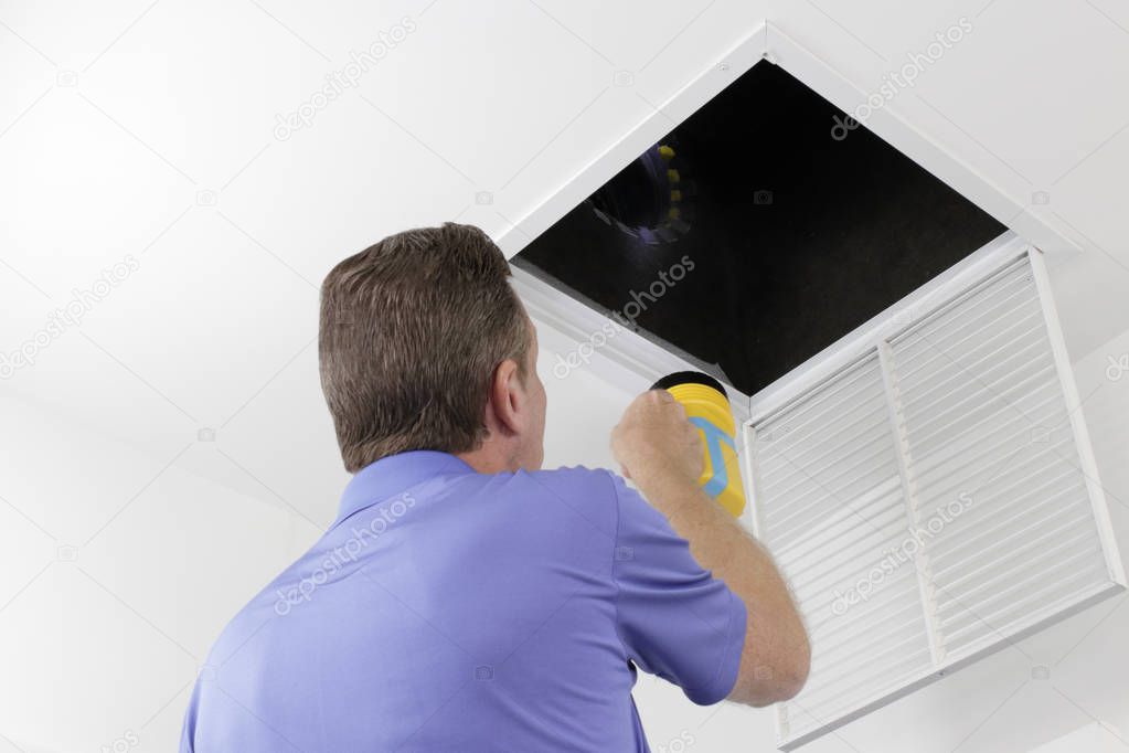 Man Inspecting an Air Duct with a Flashlight. Older male with a yellow flashlight examining HVAC ducts in a large square vent. Male technician looking over the air ducts inside a home air intake vent.