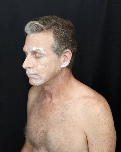 Male wearing a light bentonite clay treatment mask Thin bentonite clay face treatment worn by a guy. Thin white bentonite clay beauty treatment of mature shirtless man on black cloth curtain backgroun