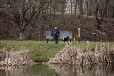 Vilnius, Lithuania - March 23, 2024: A man sits on a wooden bench with his dog near a pond in a city park, enjoying the calm early spring atmosphere.