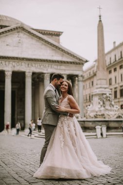 Yeni evli iki genç çekici yürüyor ve düğün günlerinde arka planda güzel ve antik mimari ile Roma'da poz