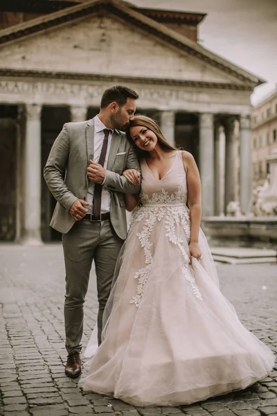 Yeni evli iki genç çekici yürüyor ve düğün günlerinde arka planda güzel ve antik mimari ile Roma'da poz