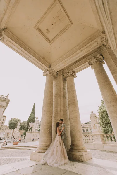 Yeni evli iki genç çekici yürüyor ve düğün günlerinde arka planda güzel ve antik mimari ile Roma'da poz