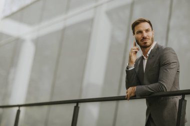 Gri takım elbise giyiyor ve onun smartphone modern ofis veya gökdelenler yanında dururken tutan genç başarılı iş adamı