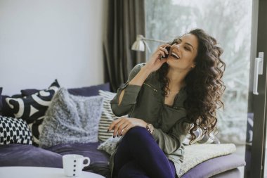 Cep telefonu ve holding kupa odasında kanepede otururken kullanan kıvırcık saçlı Genç kadın