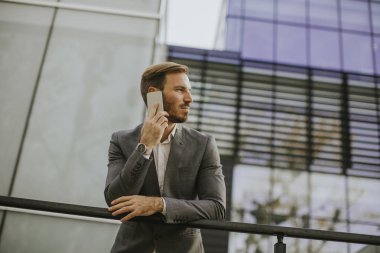 Gri takım elbise giyiyor ve onun smartphone modern ofis veya gökdelenler yanında dururken tutan genç başarılı iş adamı