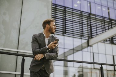 Gri takım elbise giyiyor ve onun smartphone modern ofis veya gökdelenler yanında dururken tutan genç başarılı iş adamı