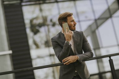 Gri takım elbise giyiyor ve onun smartphone modern ofis veya gökdelenler yanında dururken tutan genç başarılı iş adamı