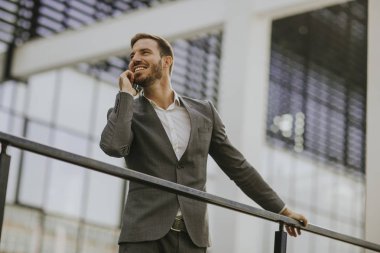 Gri takım elbise giyiyor ve onun smartphone modern ofis veya gökdelenler yanında dururken tutan genç başarılı iş adamı