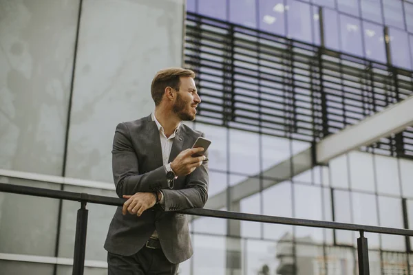 年轻的成功商人 穿着灰色西装和持有他的智能手机 同时站在现代化的办公室还是摩天大楼附近 — 图库照片