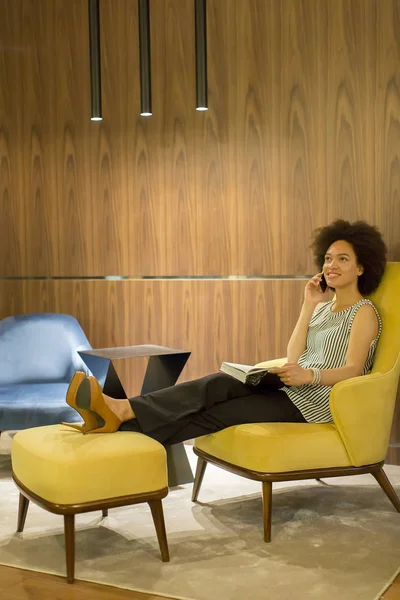 Sarı bir sandalyede oturuyor, bir cep telefonu kullanarak ve bir kitap okuma kıvırcık saçlı genç kadın