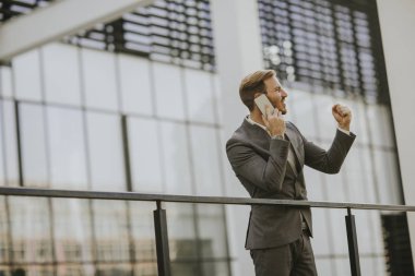 Gri takım elbise giyiyor ve onun smartphone modern ofis veya gökdelenler yanında dururken tutan genç başarılı iş adamı