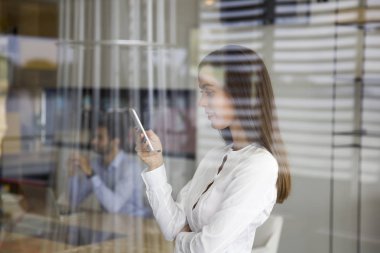 Genç ve güzel kadın cep telefonuyla cam arkasında ofiste