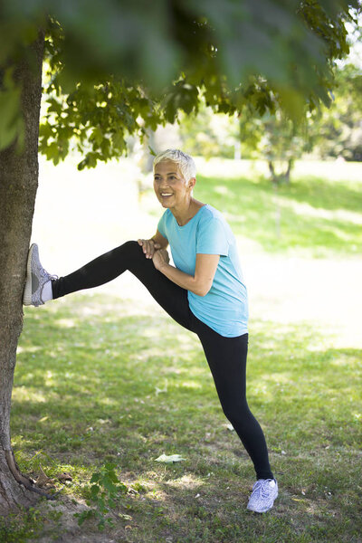 Старшая женщина делает упражнения на растяжку в парке за деревом
