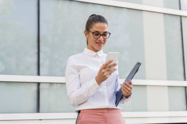 Smartphone kullanarak ve belgeler açık havada tutan başarılı genç iş kadını portresi.