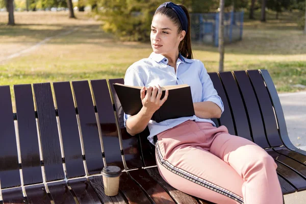 チアフル若い女性保持開いている本と秋の公園のベンチで読書 — ストック写真