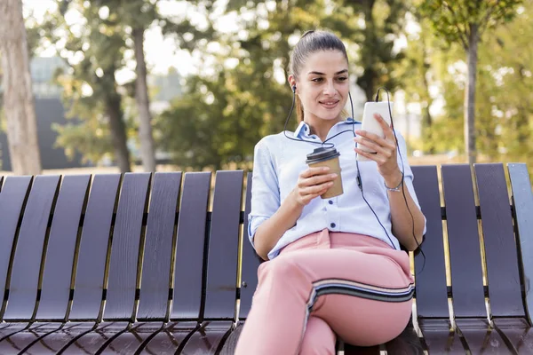 若い女性は散歩の後 秋の公園でベンチにかかっているし 晴れた日に携帯電話で音楽を聴いて — ストック写真