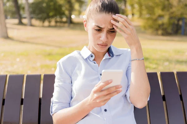 Unhappy woman sitting on bench in autumn park with mobile phone smartphone and reading sms