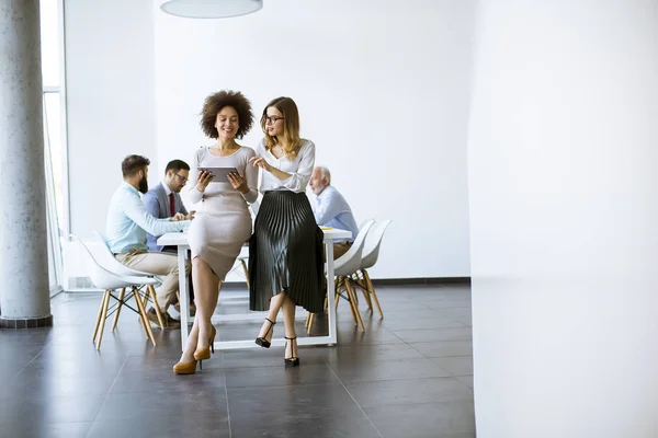 Two young businesswomen with a tablet in the office and other business people working at a desk