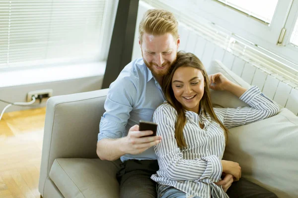カジュアルな服装 携帯電話上で何かを見ながら居間でソファに座っての笑顔のカップル — ストック写真