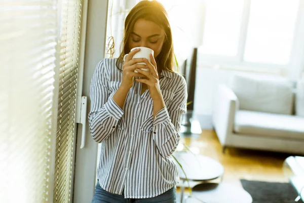 Attractive pretty young woman holding cup with hot tea or coffee and looking at the sunrise standing near the window in room