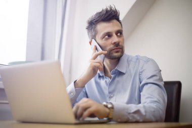 Genç ve yakışıklı işadamı cep telefonu kullanarak ve dizüstü bilgisayarda çalışma