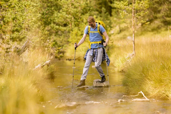 stock image Young man crosses a mountain stream during a hiking on a sunny day