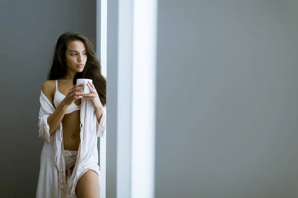 年轻的女人 穿着咖啡或茶 在房间的窗户旁边站着 — 图库照片