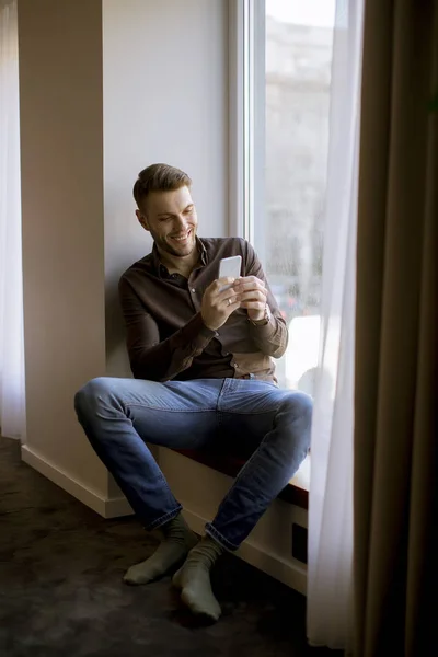 Pohledný Mladý Muž Používající Mobilní Telefon Pokoji Okna — Stock fotografie