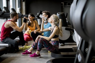 Spor giyimli bir grup genç, antrenman dan sonra spor salonunun zemininde otururken birlikte konuşup gülüyor